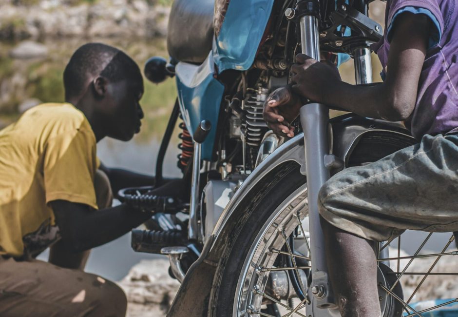 Two young African boys clean a motorcycle outdoors, showcasing teamwork and fun on a sunny day.